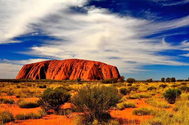 daytime image of orange Ayers rock with dry bushland in foreground and wispy clouds in the sky