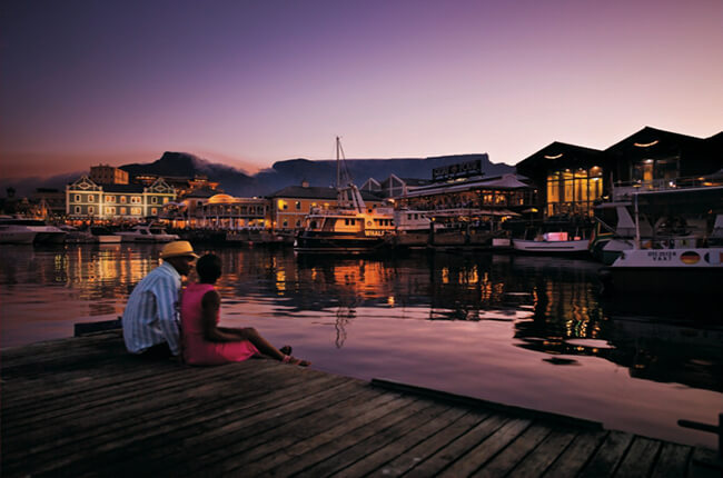 Couple sitting together at the end of a pier in Cape Town Bay, with docked boats and waterfront restaurants in the distance