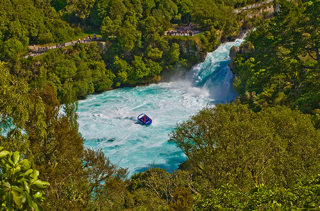 The emerald waters of Huka Falls surrounded by the green vegetation of the forest. Shotover Jet in the centre of the plunge pool