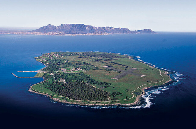 Aerial view of Robben Island, surrounded by deep blue ocean, and Cape Point far away in the background.