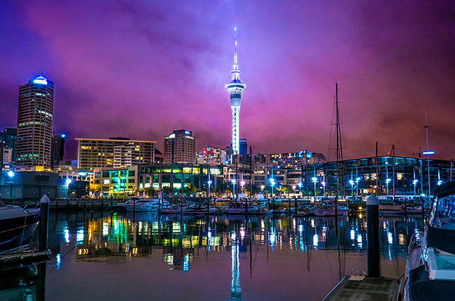 Auckland Harbour lit up at night time, with the Sky Tower dominating the vibrant purple skies