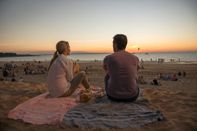 Couple sat on Mindil Beach in Darwin at sunset with many people on the beach in front of them