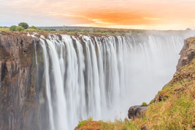 Victoria Falls with water flowing over the falls and mist, with orange sky