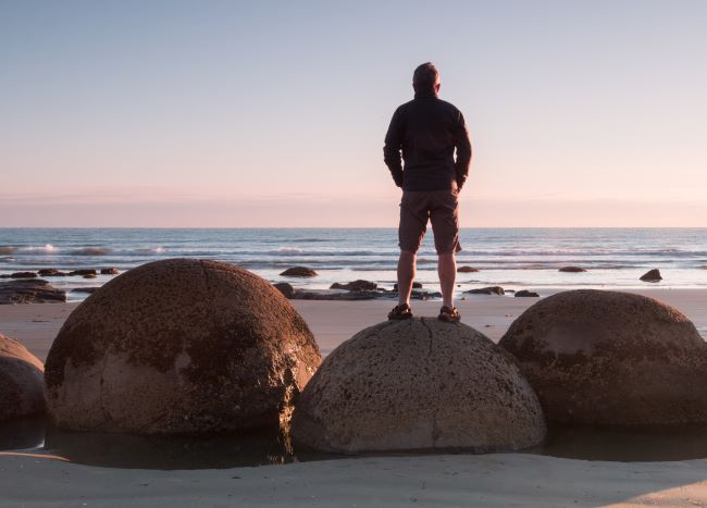 Man standing on the large Moeraki Boulders, with the calm waves of the water in the background