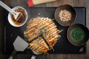 Okonomiyaki being made with condiments around the side